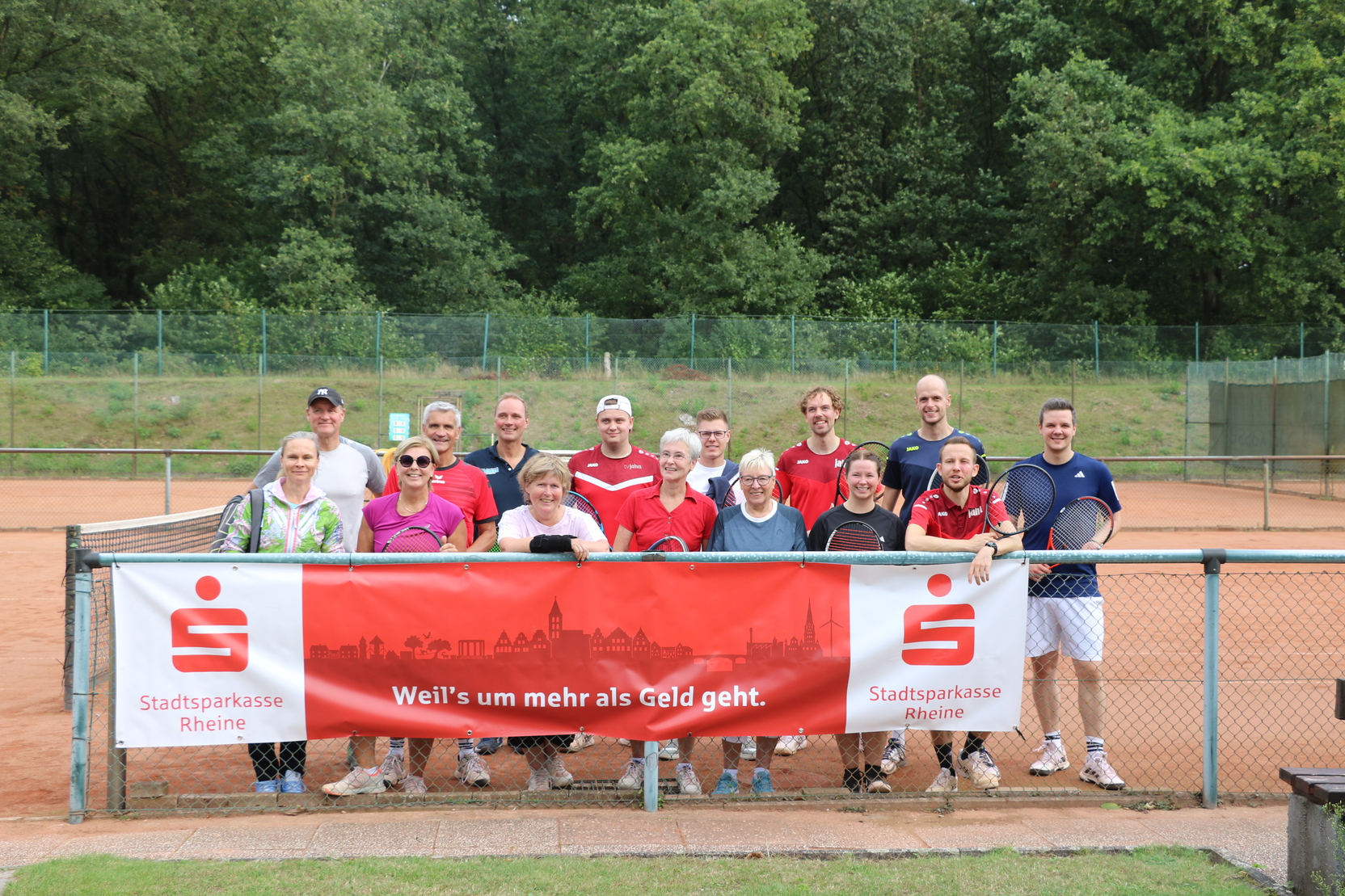 Gruppeneinstellung von 16 Personen auf einem Tennisplatz, mit einem Banner der Stadtsparkasse Rheine im Vordergrund.