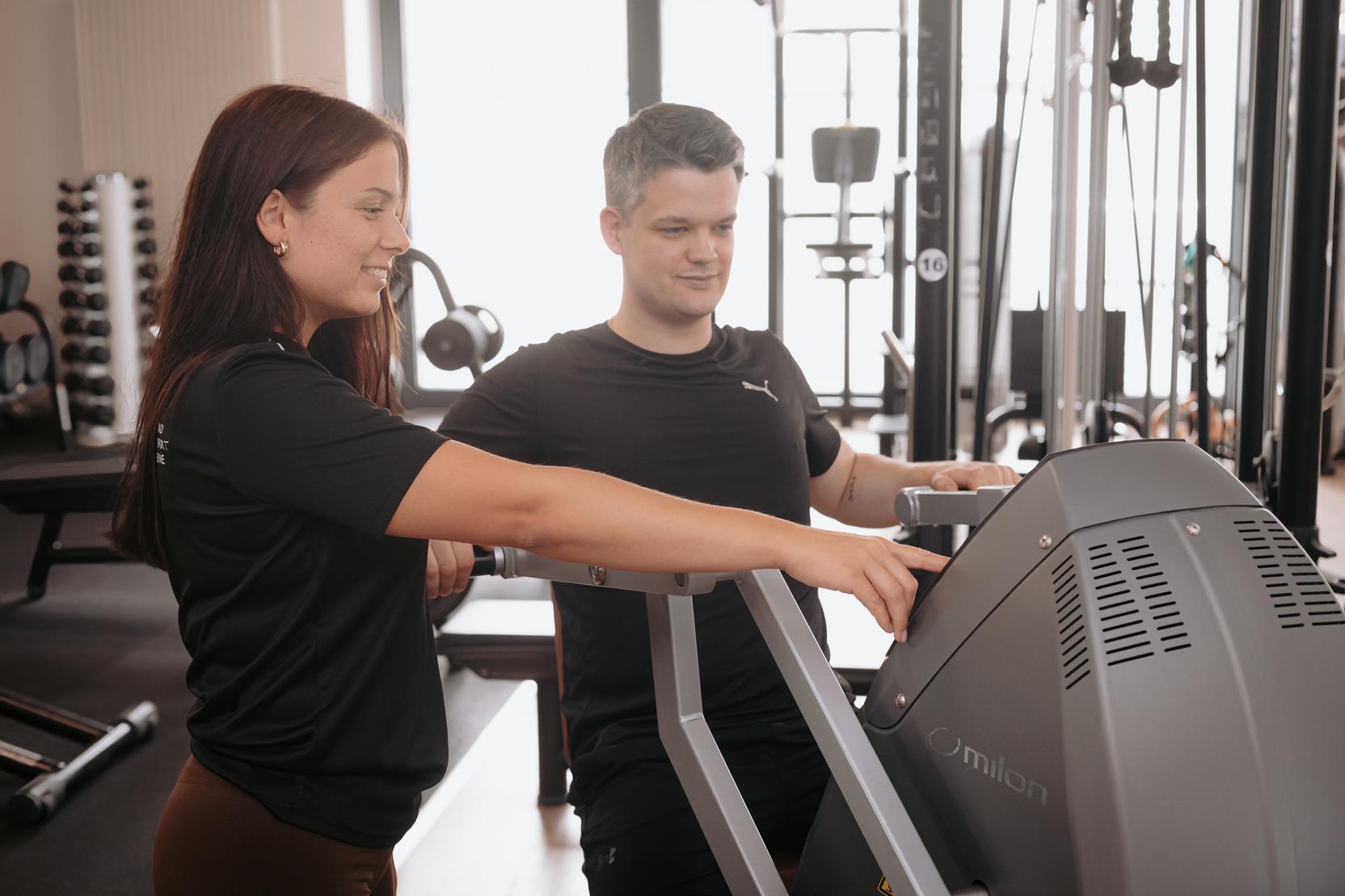Trainerin erkl&auml;rt einem Mann die Bedienung eines Fitnessger&auml;ts in einem modernen Fitnessstudio.