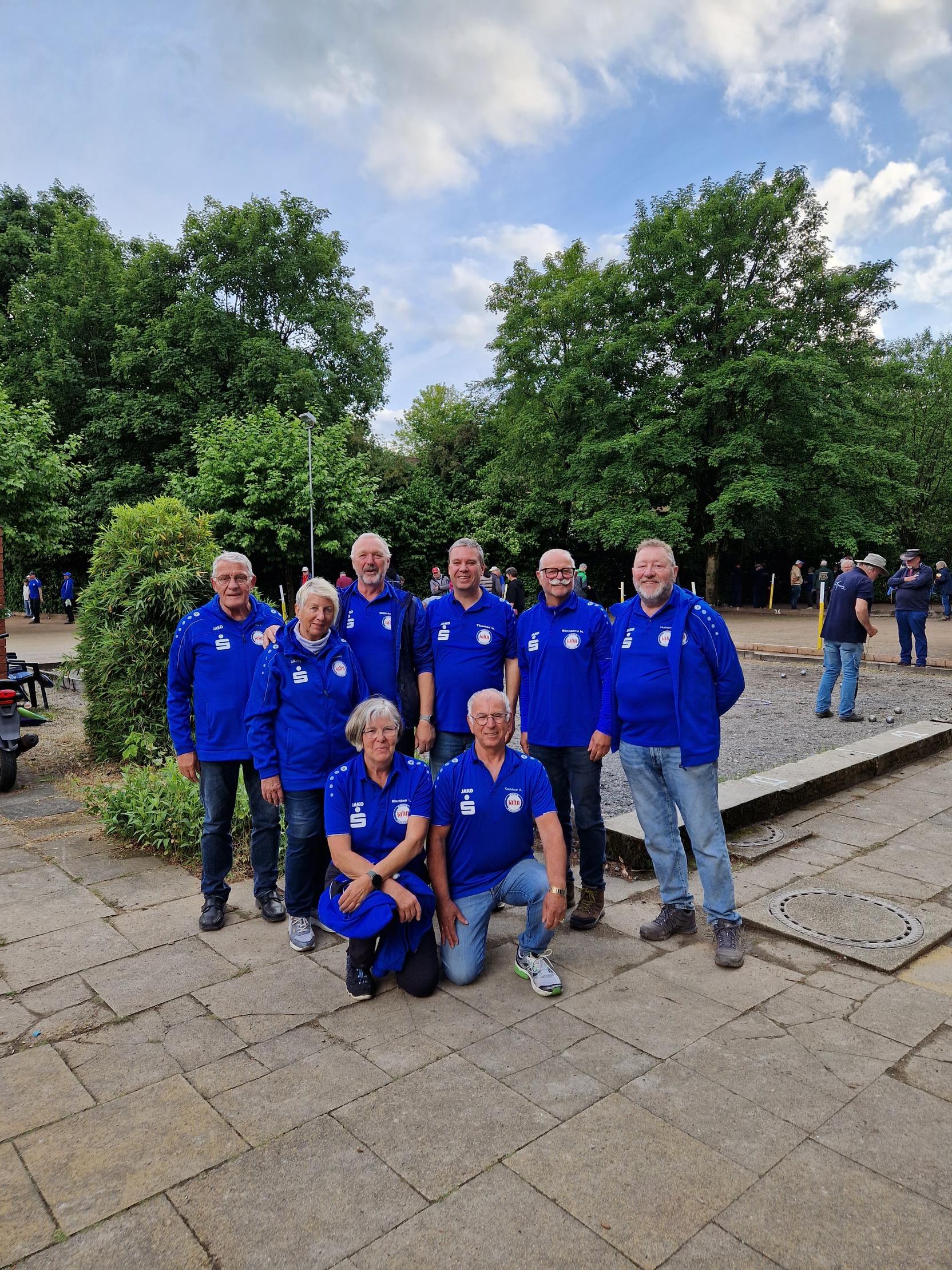 Gruppe von acht Personen in blauen Trikots, stehend und sitzend, auf einem Platz mit Bäumen im Hintergrund.