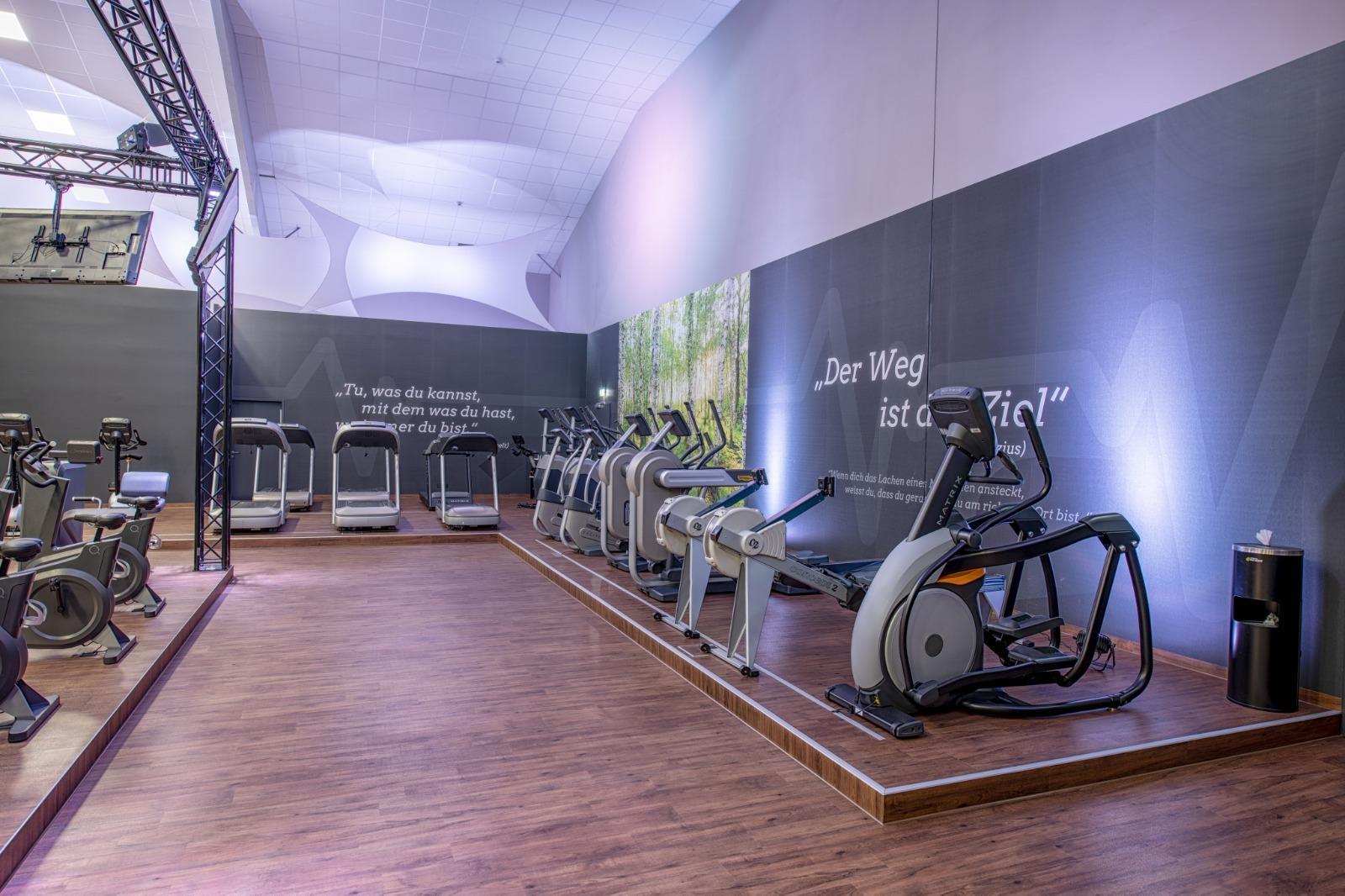 Fitnessstudio mit mehreren Cardioger&auml;ten und motivationalem Zitat an der Wand, Holzfu&szlig;boden und sanfte Beleuchtung.