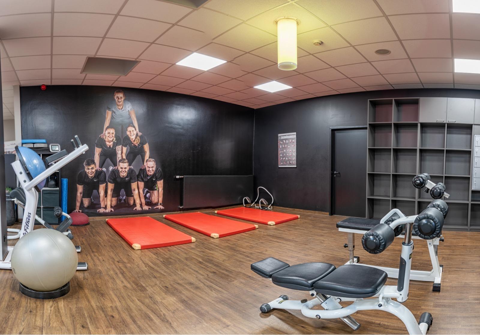 Fitnessraum mit Matten, Hanteln und einem Fitnessger&auml;t, im Hintergrund ein Wandbild von mehreren Sportlern.