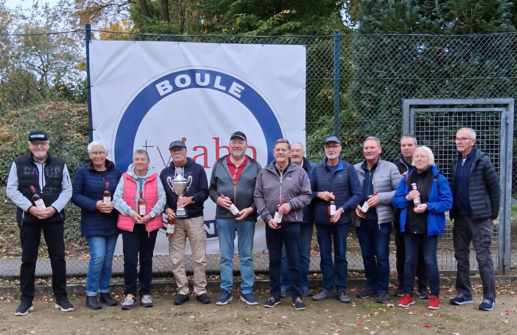 Gruppe von zwölf Personen mit Pokal und Flaschen vor Boule-Plakat auf einem Freiluftplatz.