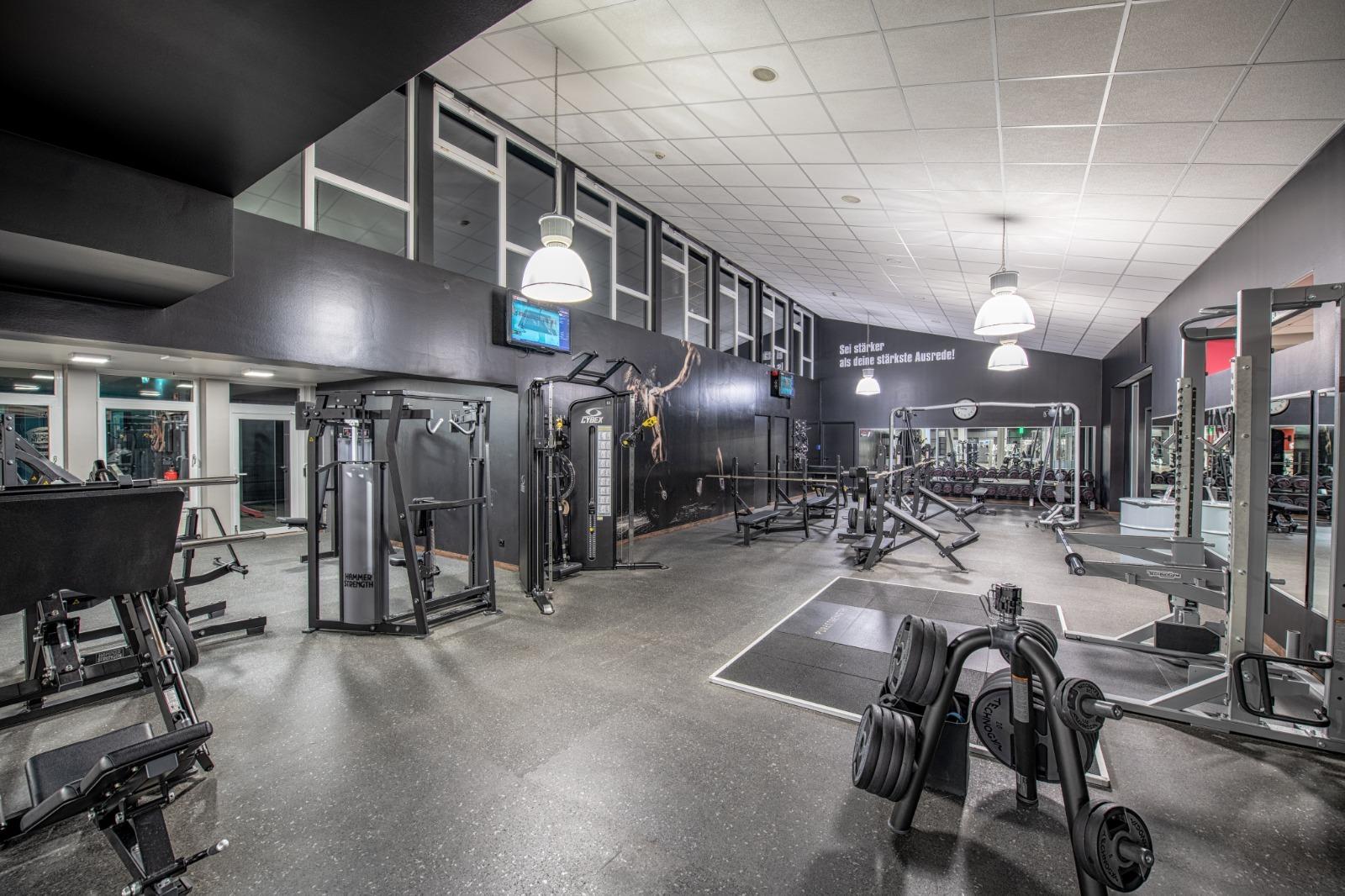 Modernes Fitnessstudio mit verschiedenen Trainingsger&auml;ten, Hanteln und gro&szlig;en Fenstern, in schlichten dunklen Farben gestaltet.