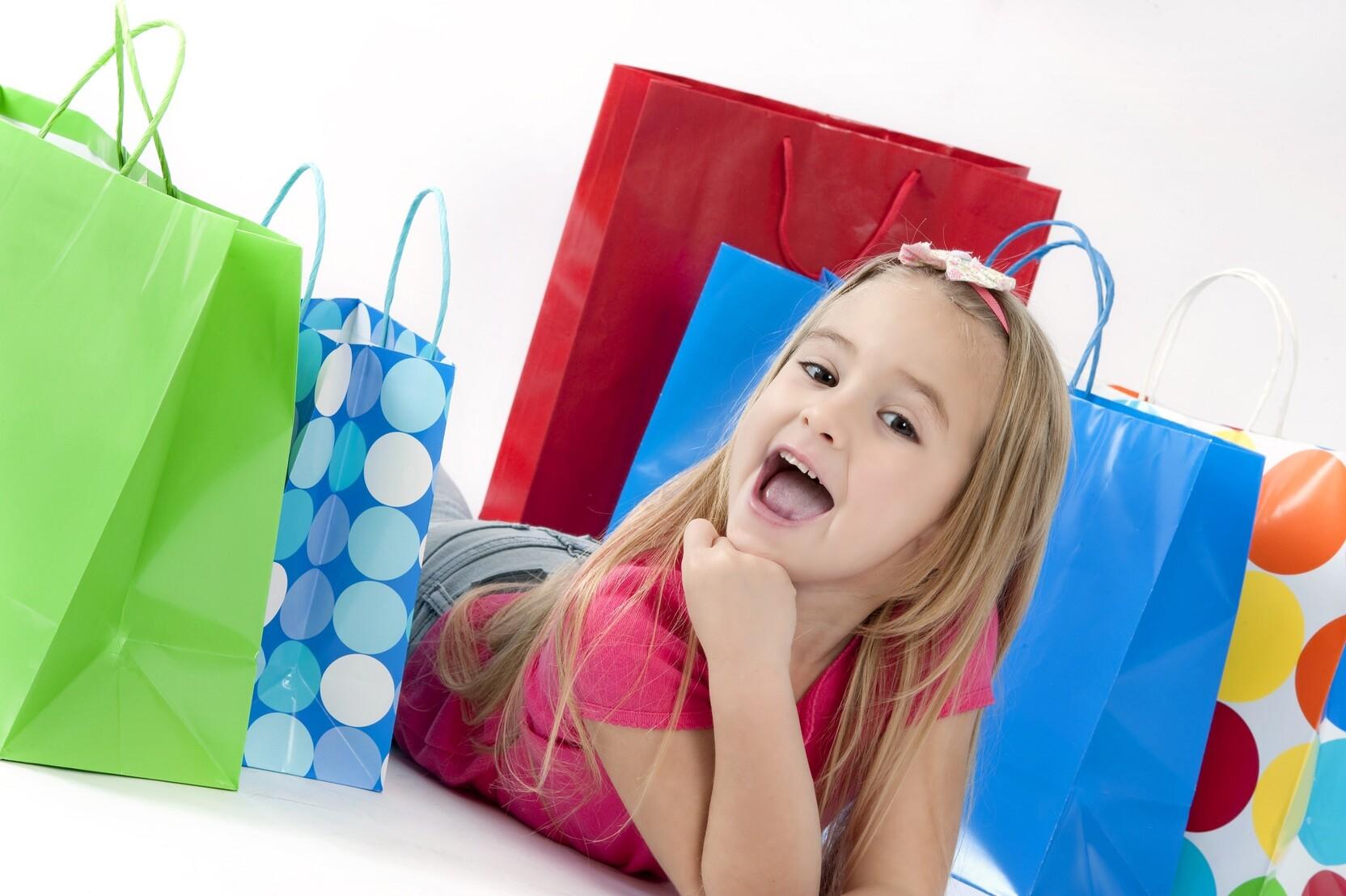L&auml;chelndes M&auml;dchen liegt vor bunten Einkaufstaschen in Gr&uuml;n, Blau und Rot auf einer neutralen Fl&auml;che.