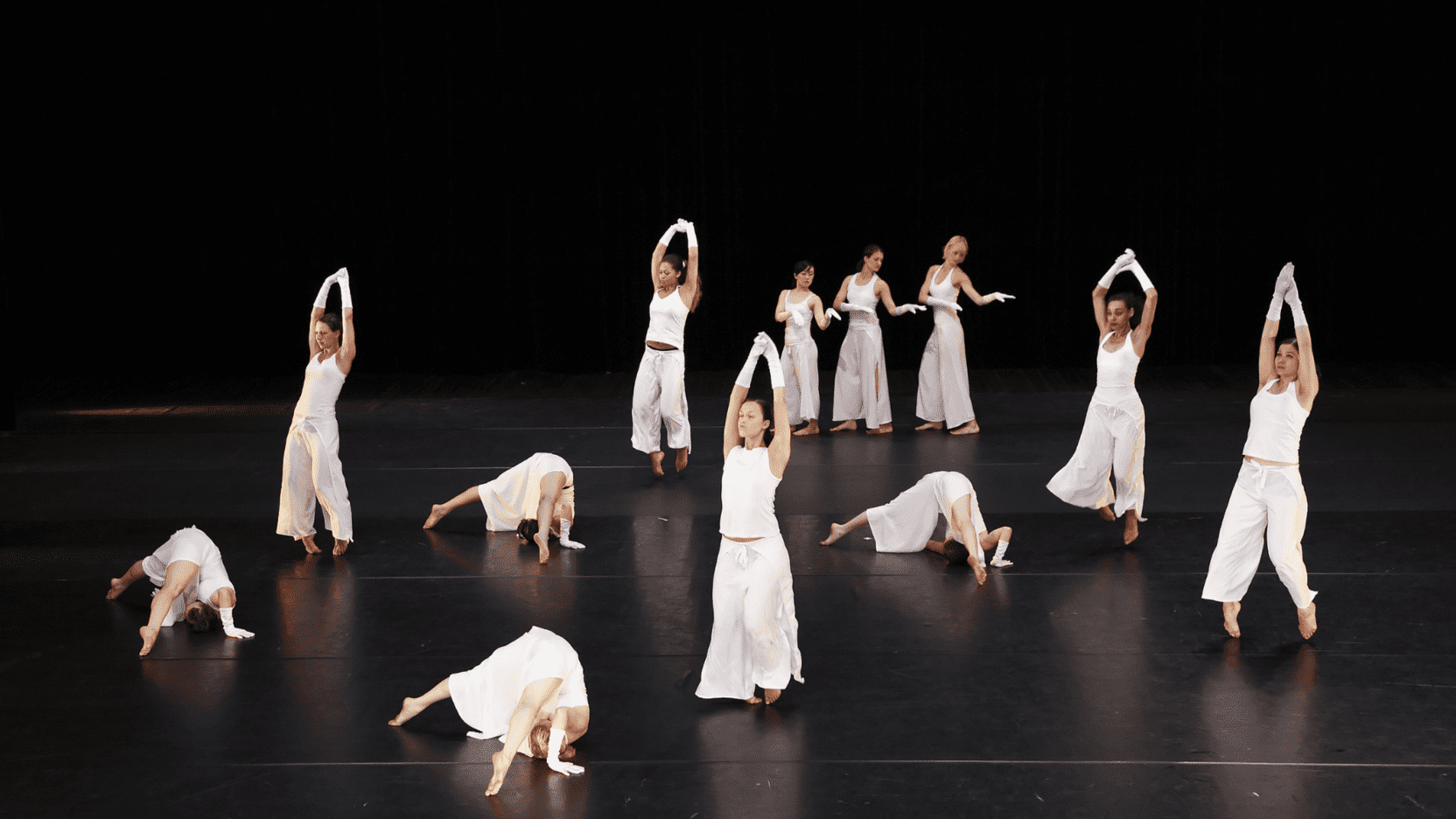 Eine Gruppe von T&auml;nzerinnen in wei&szlig;en Outfits f&uuml;hrt synchron Bewegung auf einer dunklen B&uuml;hne aus.