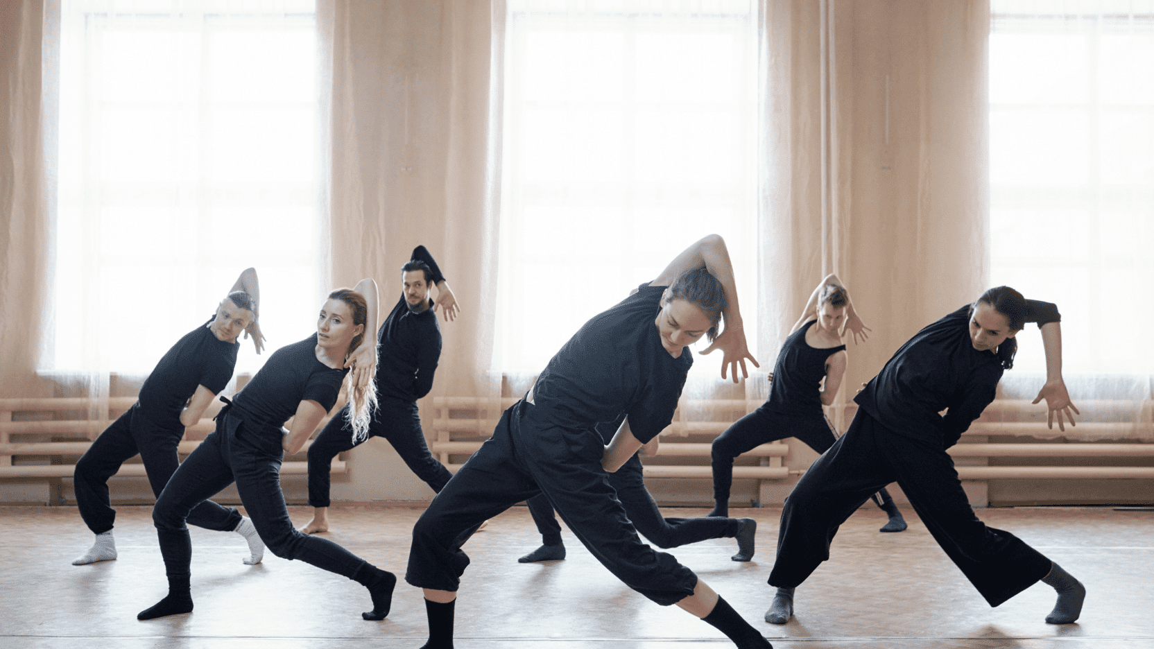Sechs T&auml;nzerinnen in schwarzer Kleidung f&uuml;hren eine zeitgen&ouml;ssische Choreografie in einem hellen Tanzstudio auf.