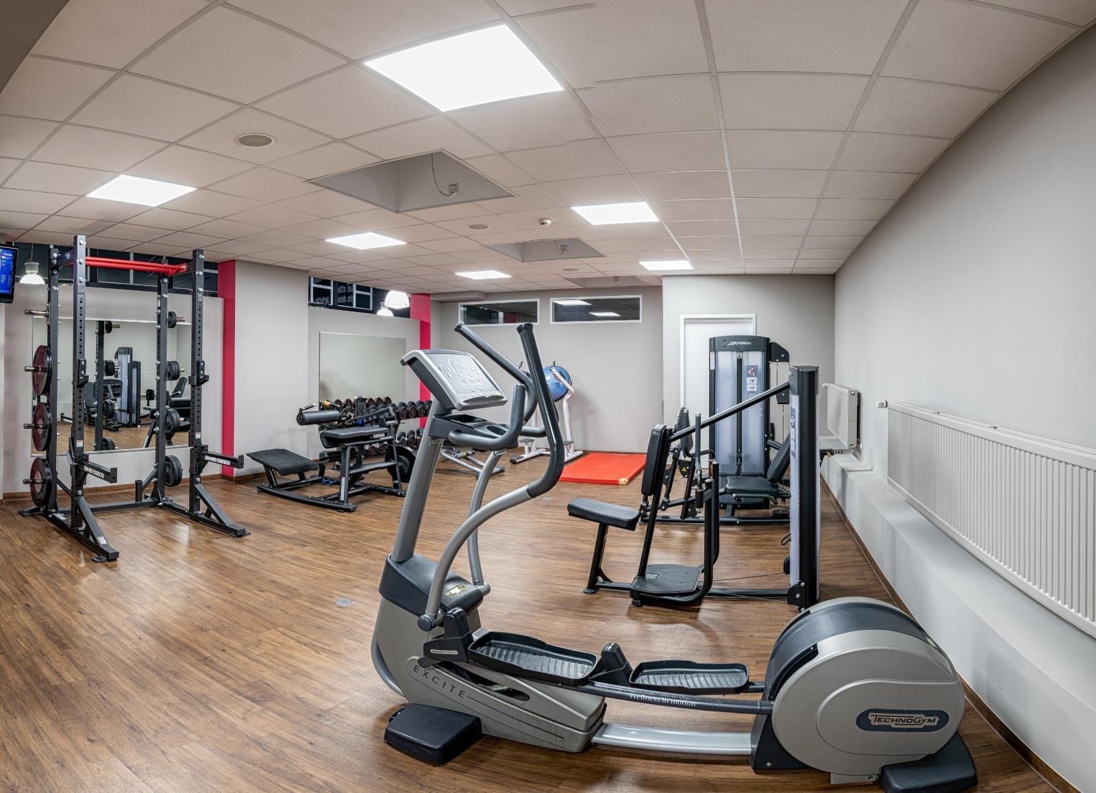 Fitnessraum mit einem Crosstrainer, Gewichten und Hanteln auf Holzfu&szlig;boden sowie Spiegelwand im Hintergrund.