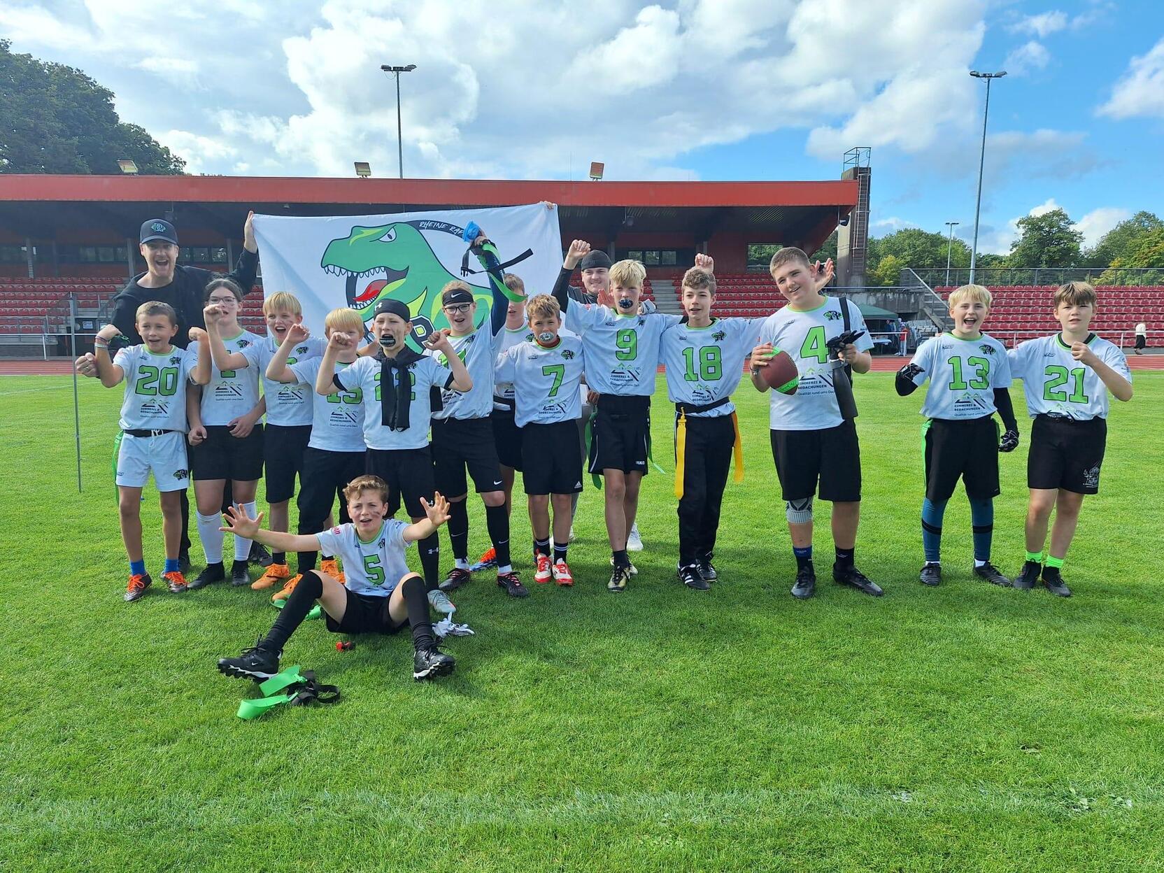 Gruppe von Jugendlichen in Sportkleidung posiert vor einer Flagge auf einem Fu&szlig;ballplatz, einige halten B&auml;lle in der Hand.