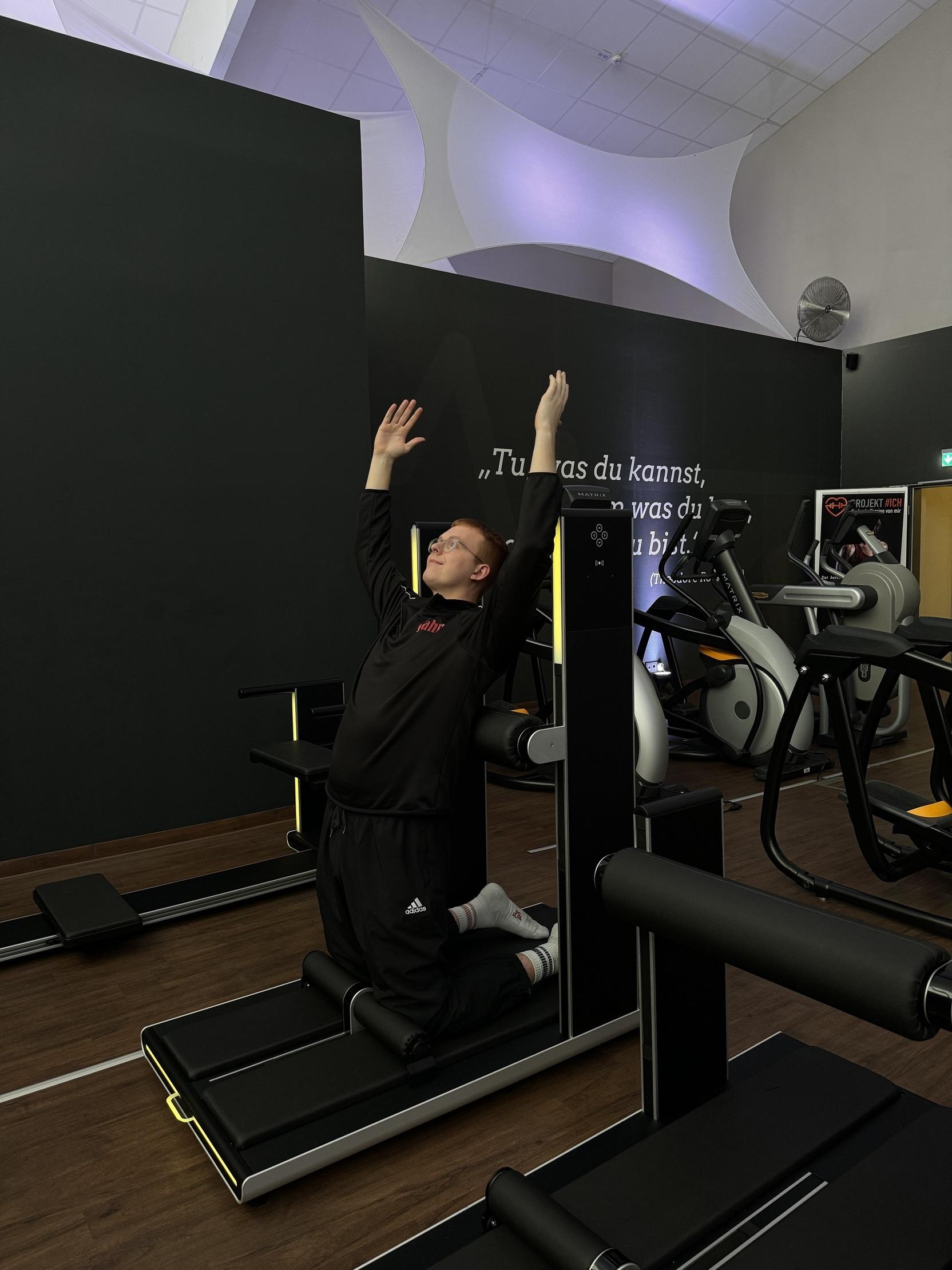 Junger Mann in Fitnesskleidung dehnt sich auf einem Trainingsgerät im Fitnessstudio. Hintergrund ist dunkel mit Geräten.