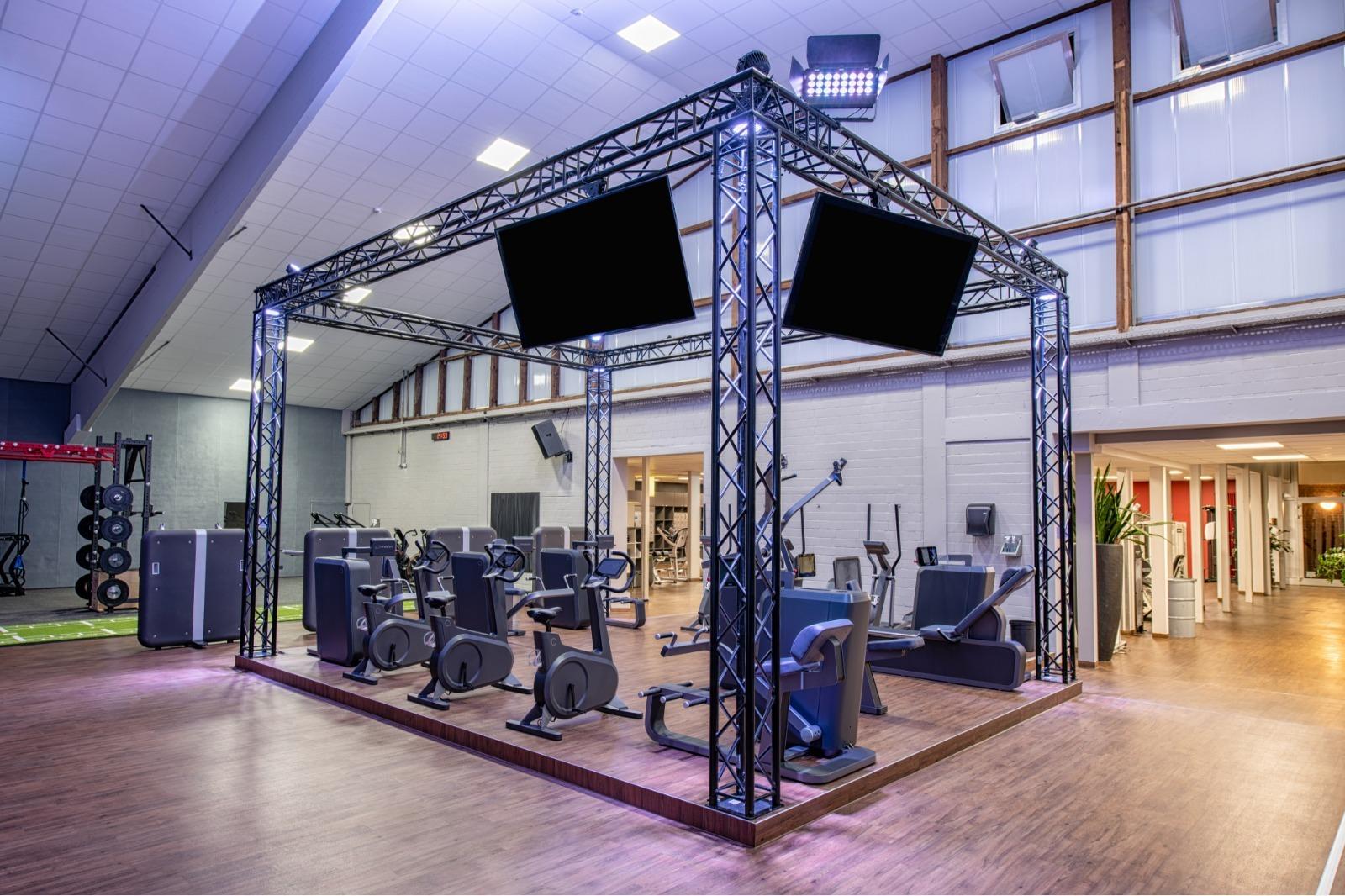 Fitnessbereich mit modernen Ger&auml;ten im Rahmen aus Metall, darunter zwei gro&szlig;e Bildschirme. Holzfu&szlig;boden und Fenster im Hintergrund.