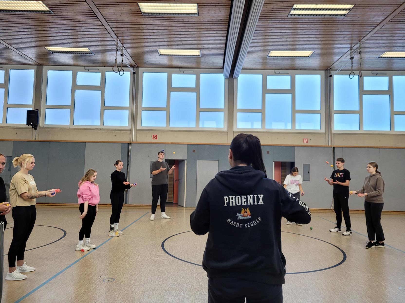 Gruppe von trainierenden Personen in einem Raum, angeleitet von einer Trainerin mit &bdquo;PHOENIX&ldquo;-Hoodie.