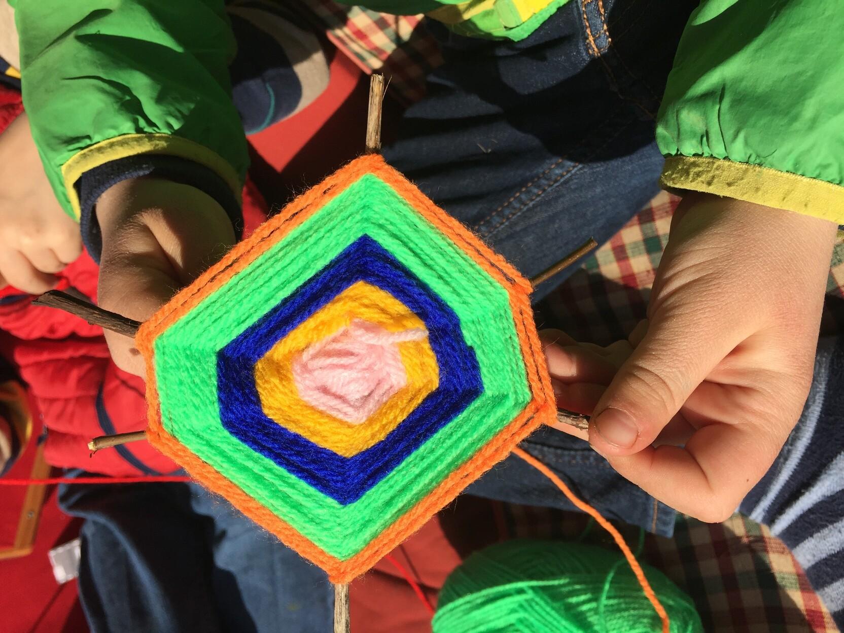 Nahaufnahme von H&auml;nden, die ein buntes, gesticktes Hexagon aus Wolle halten, vor einem karierten Untergrund.