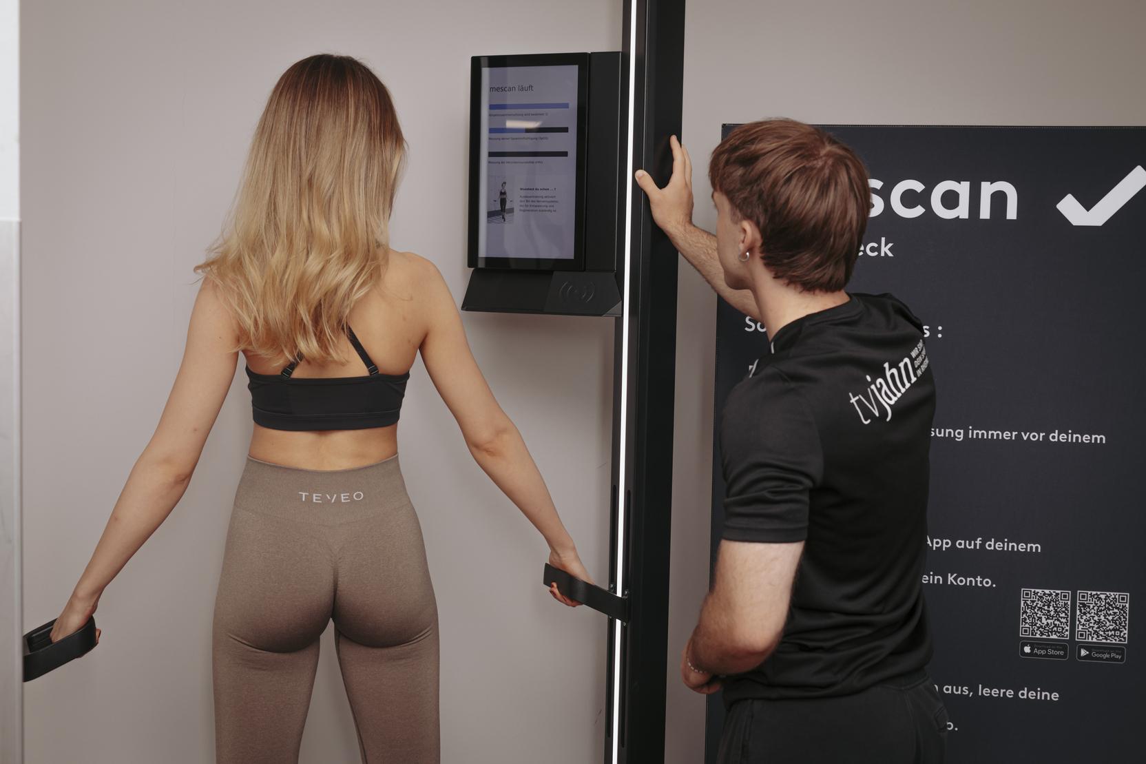 Frau in Sportbekleidung f&uuml;hrt K&ouml;rperanalyse mit Trainer an einem Ger&auml;t durch, w&auml;hrend ein Bildschirm Informationen anzeigt.