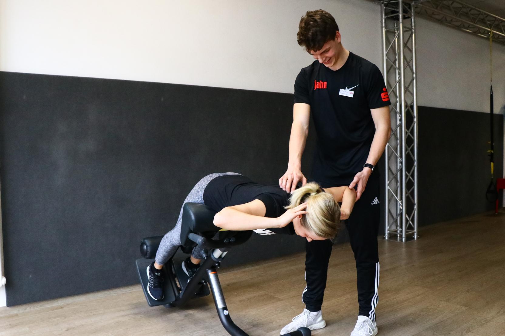 Trainer unterst&uuml;tzt Frau bei R&uuml;cken&uuml;bung auf einem Fitnessger&auml;t in einem modernen Trainingsraum.