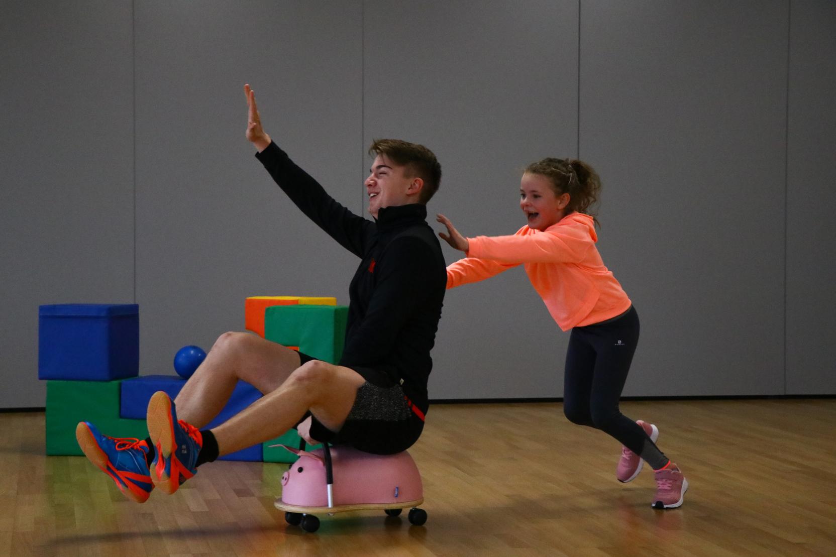 Junge sitzt auf einem Spielzeug in der Sporthalle, w&auml;hrend ein M&auml;dchen ihn anschiebt und beide lachen.