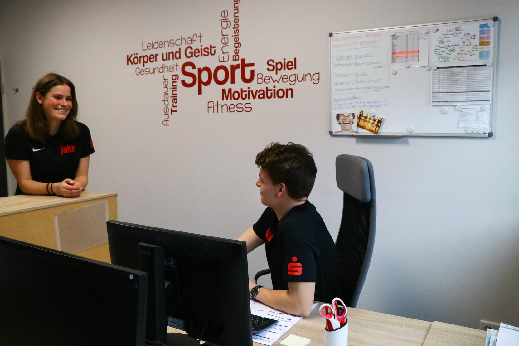 Zwei Personen in einem B&uuml;ro, eine am Empfang und eine sitzend am Tisch, umgeben von sportlichen Begriffen an der Wand.