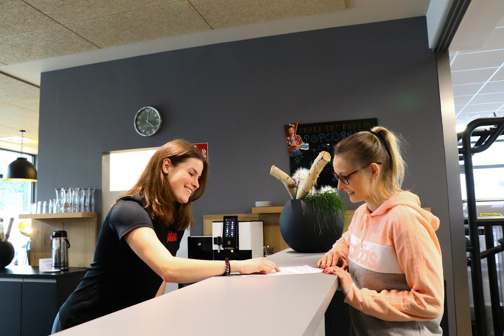 Frau am Empfangstresen &uuml;bergibt Dokument an eine Kundin in sportlicher Kleidung. Heller, moderner Empfangsbereich.