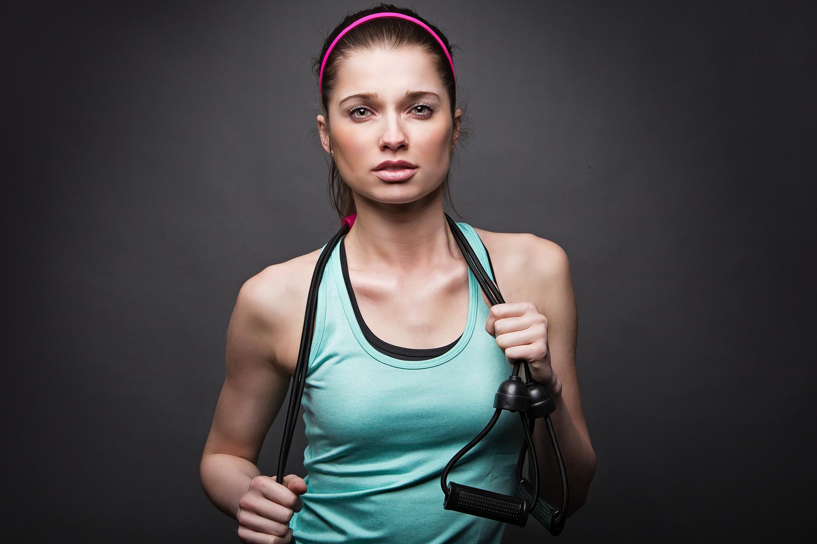 Junge Frau in Sportkleidung hält ein Widerstandsband und blickt ernst in die Kamera vor einem dunklen Hintergrund.