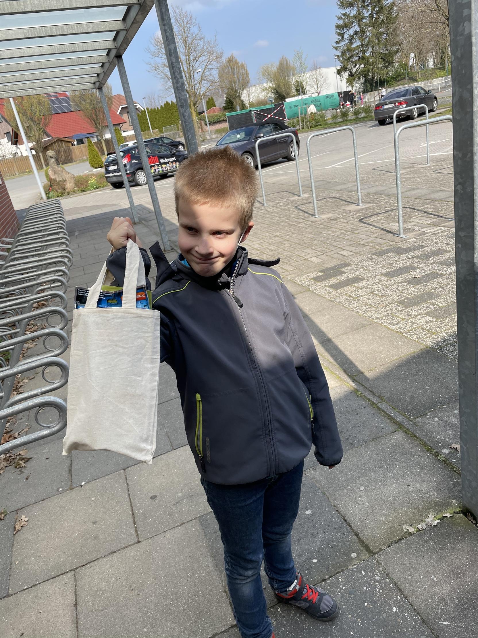 Junge h&auml;lt eine wiederverwendbare Tasche mit Getr&auml;nken auf einem Gehweg, im Hintergrund Parkpl&auml;tze und B&auml;ume.
