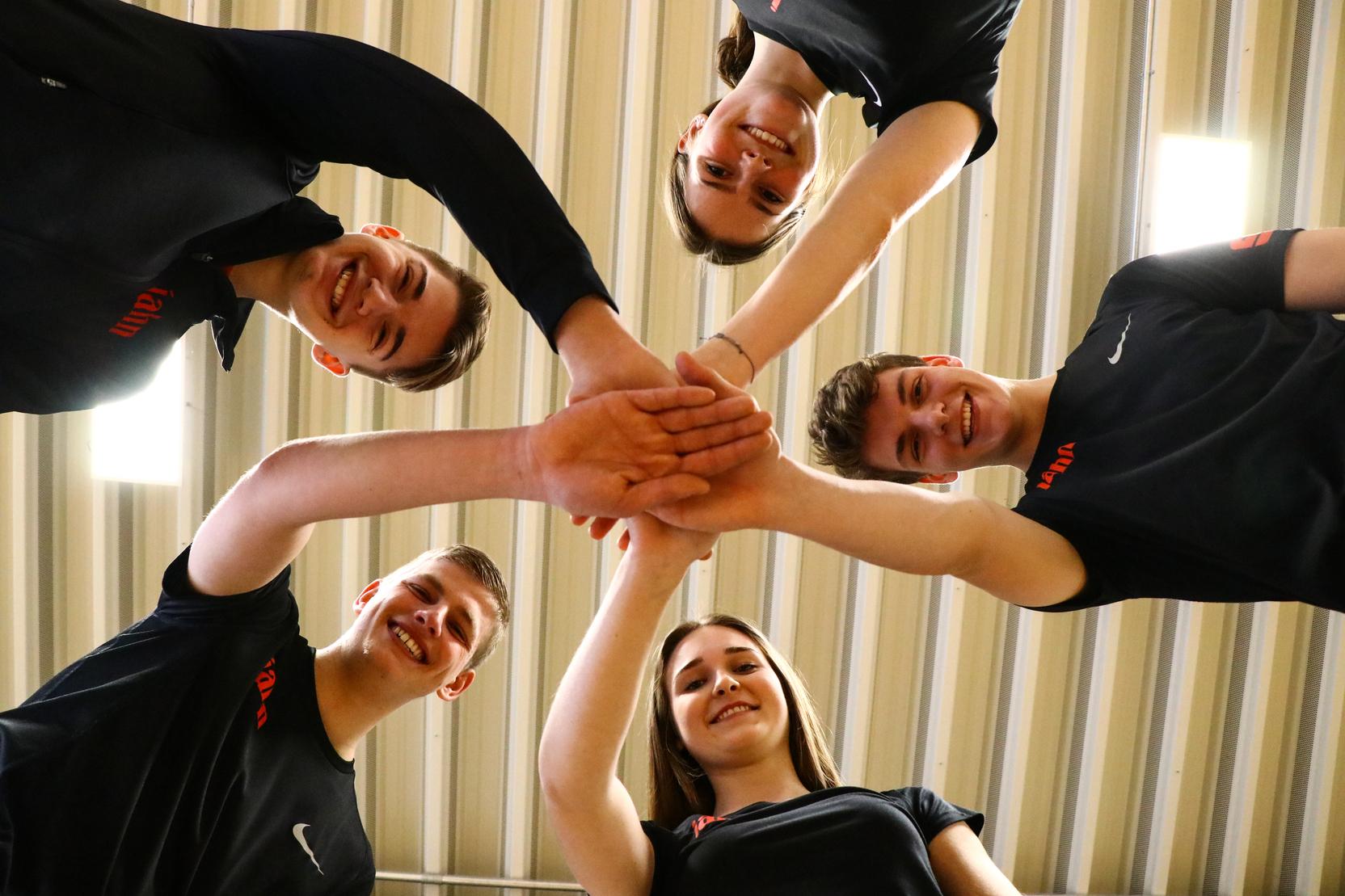 F&uuml;nf Jugendliche stehen im Kreis, legen ihre H&auml;nde zusammen und blicken nach unten auf die H&auml;nde.