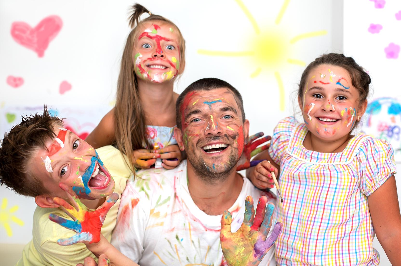Ein Mann und drei Kinder mit bemalten Gesichtern und H&auml;nden posieren fr&ouml;hlich vor einer bunten Wand.