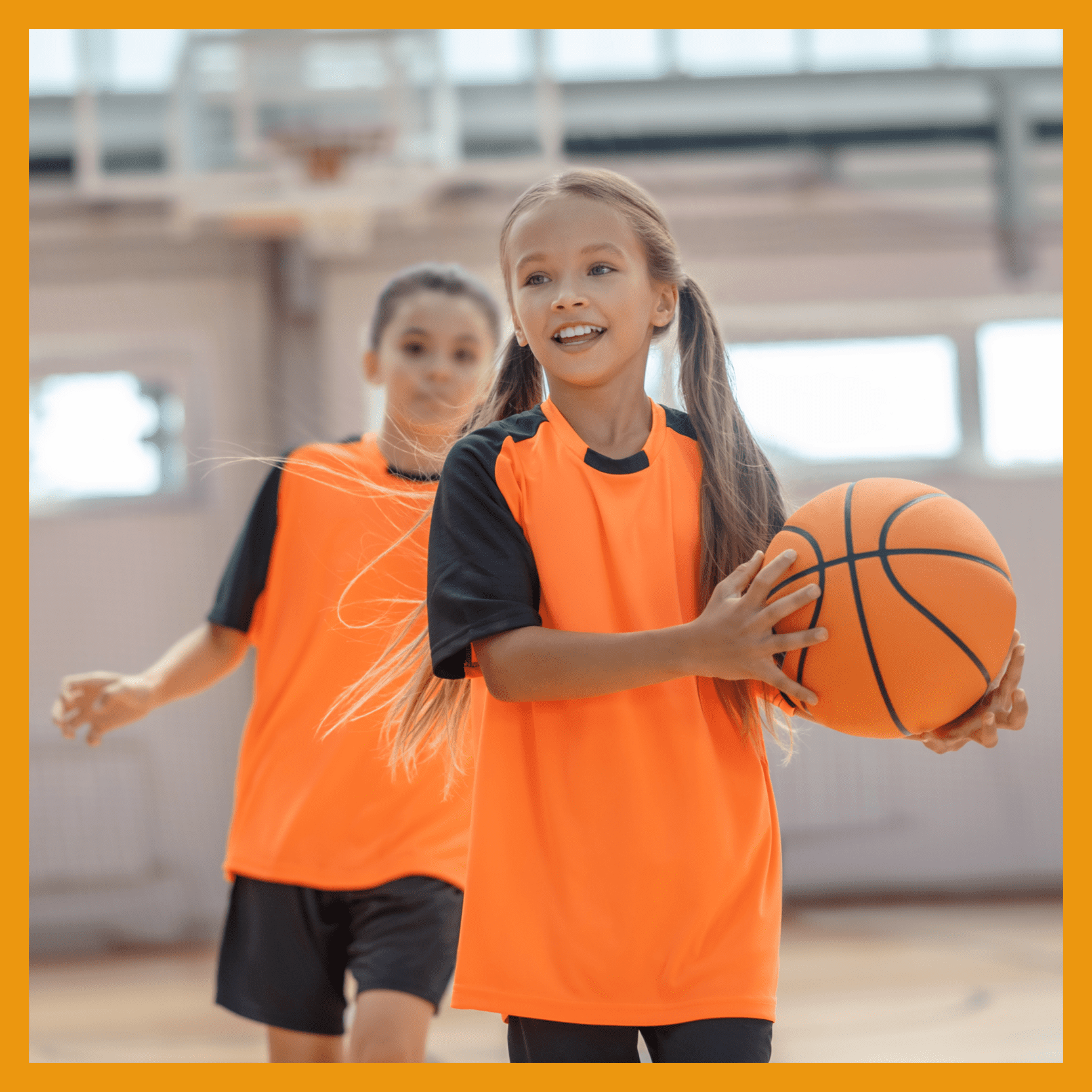 Zwei Mädchen in orangefarbenen Sporttrikots spielen Basketball in einer Turnhalle. Eines trägt den Ball, das andere läuft daneben.