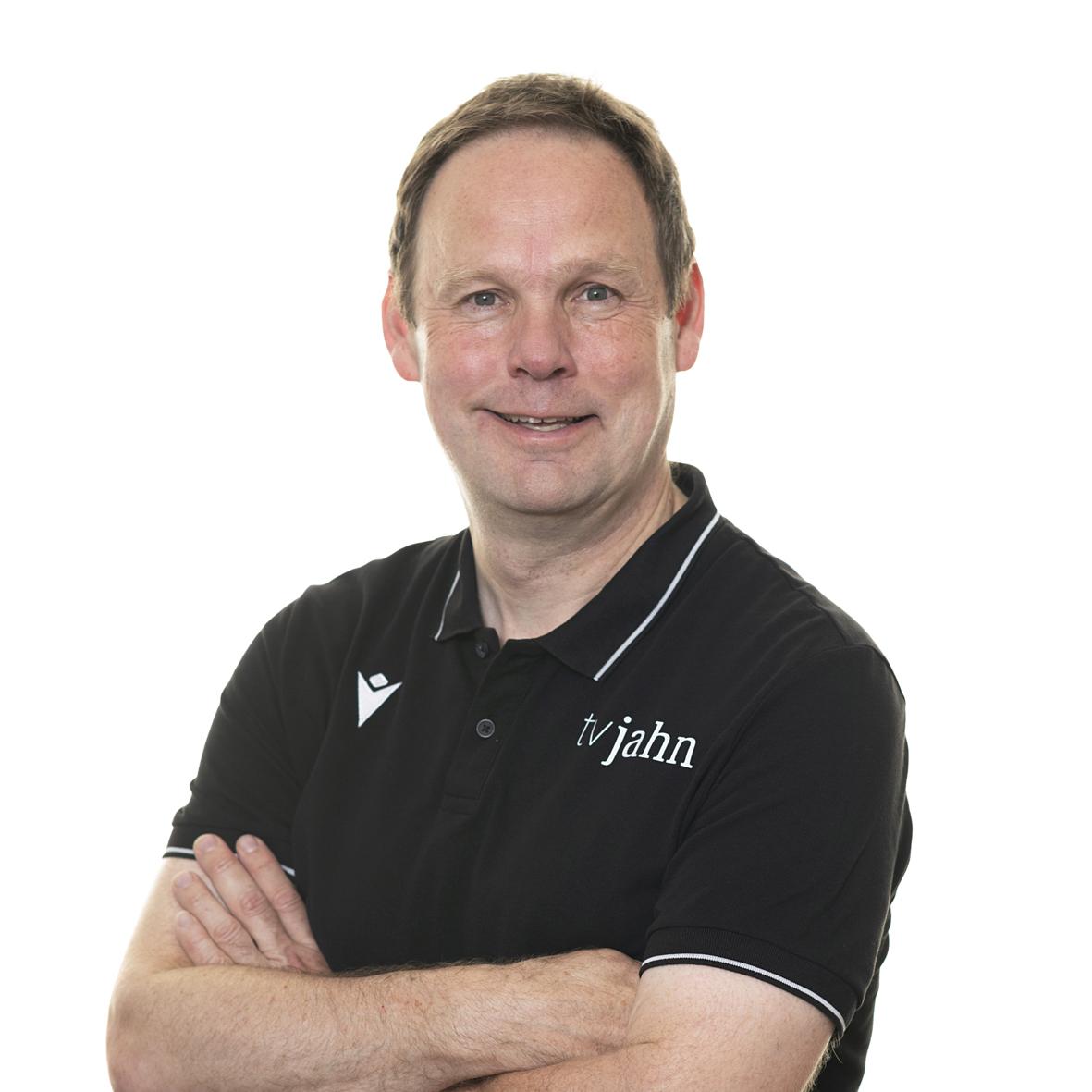 Mann mit kurzen Haaren und lächelndem Gesicht, trägt ein schwarzes Polohemd mit Aufschrift „tv jahn“, Arme verschränkt.
