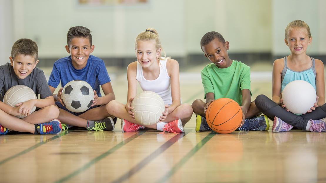 F&uuml;nf Kinder sitzen auf einem Sportboden und halten verschiedene B&auml;lle: Fu&szlig;ball, Volleyball, Basketball und mehr.