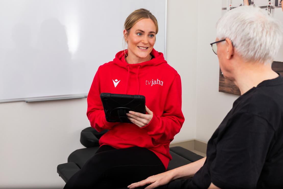 Therapeutin in rotem Hoodie sitzt mit Tablet vor einem &auml;lteren Mann, beide in einem hellen Beratungsraum.