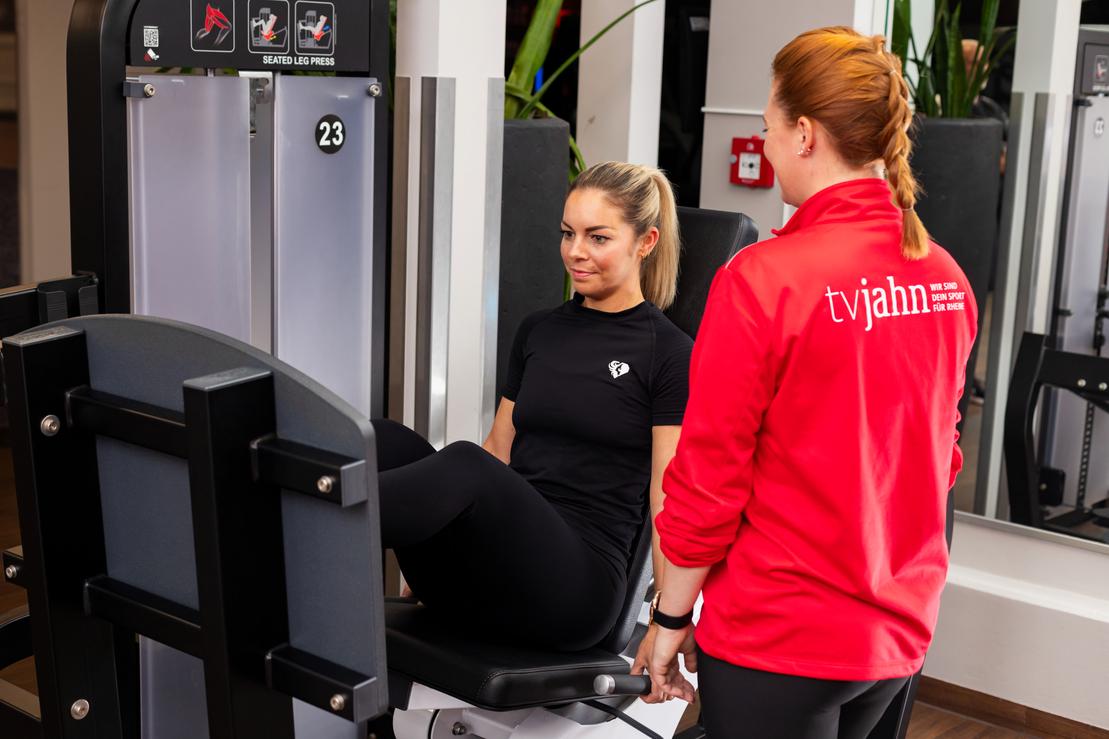 Frau trainiert am Beinstrecker-Ger&auml;t unter Anleitung einer Trainerin im Fitnessstudio.