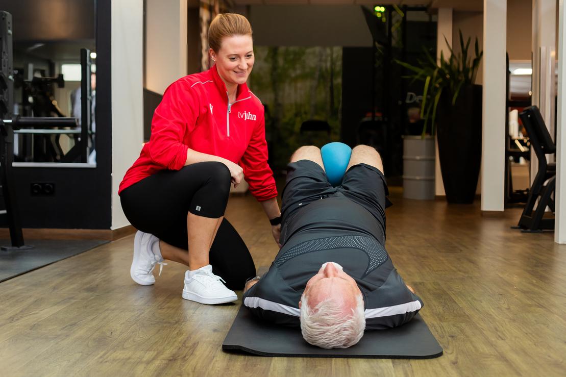 Fitnesscoach unterst&uuml;tzt m&auml;nnlichen Klienten bei einer &Uuml;bung auf einer Matte in einem modernen Fitnessstudio.