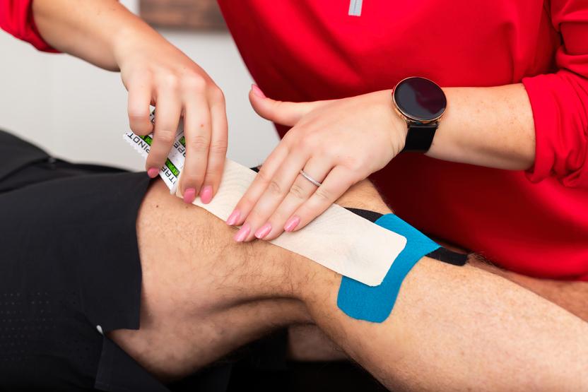 Eine Person klebt ein Verbandst&uuml;ck auf das Bein eines anderen, w&auml;hrend sie eine Tubenbandage in der Hand h&auml;lt.