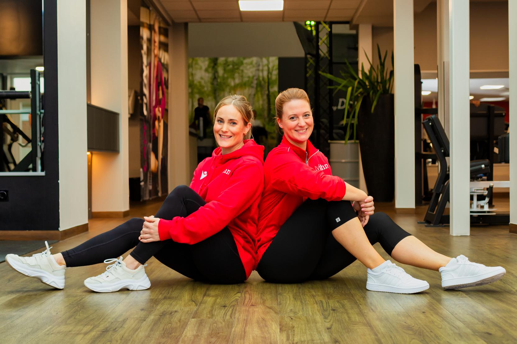 Zwei Frauen sitzen in Sportkleidung R&uuml;cken an R&uuml;cken auf dem Boden eines Fitnessstudios.