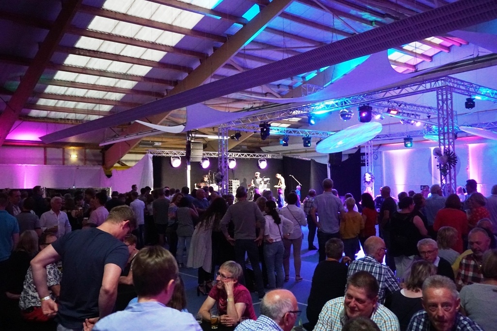 Blick auf eine Menschenmenge bei einem Konzert in einer beleuchteten Halle, B&uuml;hne im Hintergrund sichtbar.