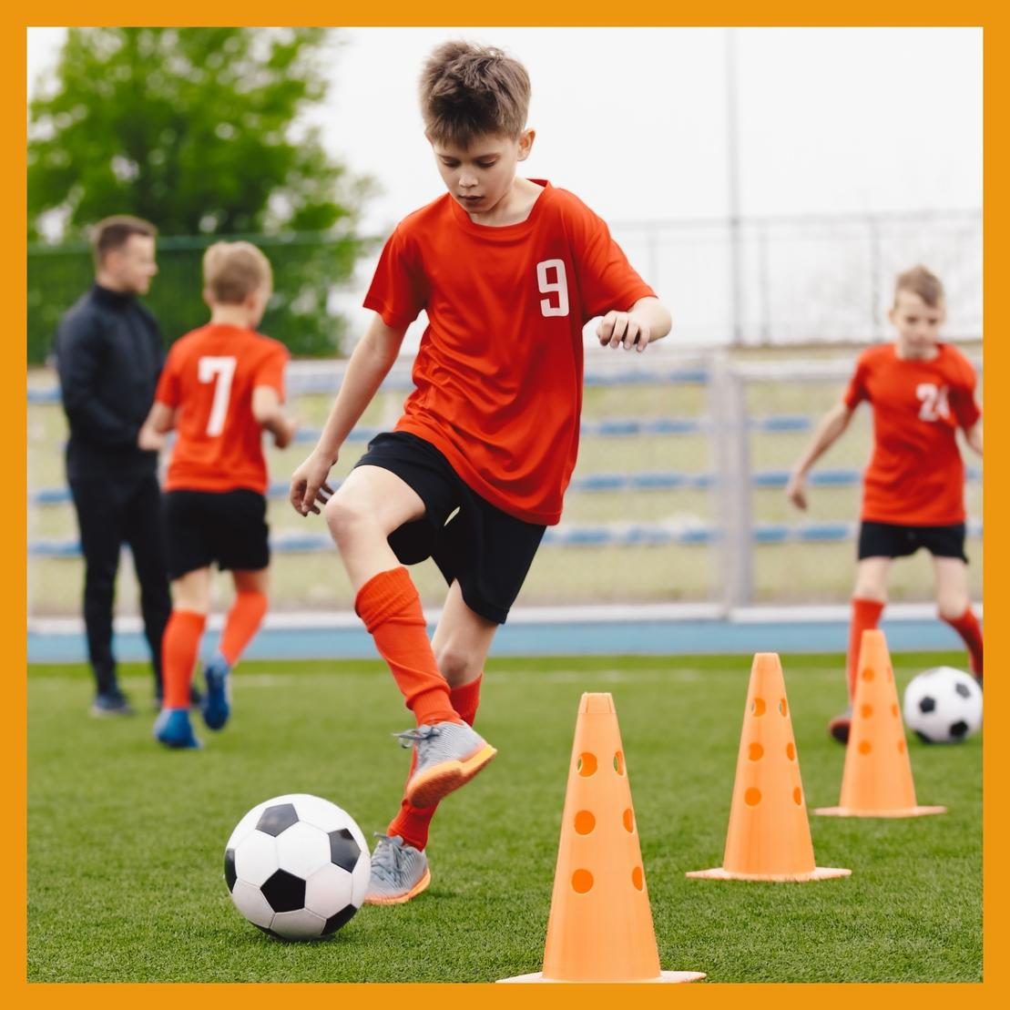 Junge in rotem Trikot dribbelt einen Fu&szlig;ball zwischen orangefarbenen H&uuml;tchen auf einem Kunstrasenplatz.