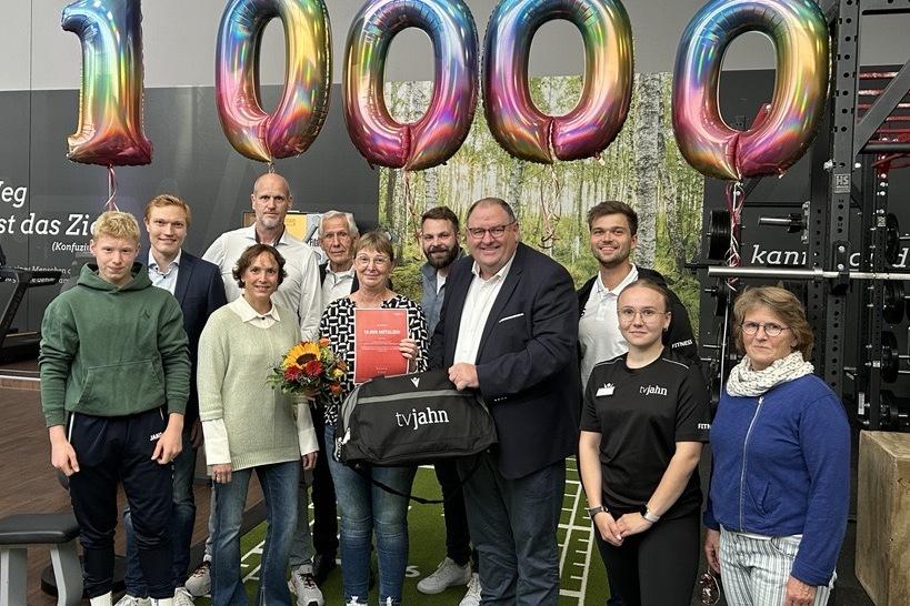Gruppe von Personen feiert Jubil&auml;um mit &bdquo;1000&ldquo;-Luftballons in einem Fitnessstudio, Blumenstrau&szlig; und Geschenkt&uuml;te in der Mitte.