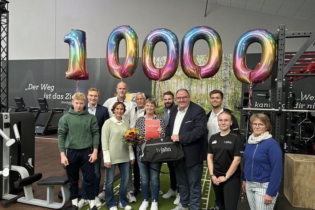 Gruppe von Personen feiert Jubil&auml;um mit &bdquo;1000&ldquo;-Luftballons in einem Fitnessstudio, Blumenstrau&szlig; und Geschenkt&uuml;te in der Mitte.
