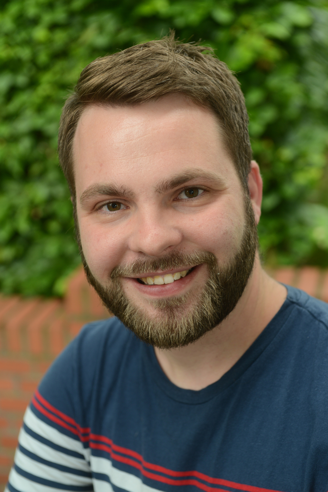 L&auml;chelnder Mann mit Bart in T-Shirt vor gr&uuml;ner Hecke, auf einem Mauersteinhintergrund.