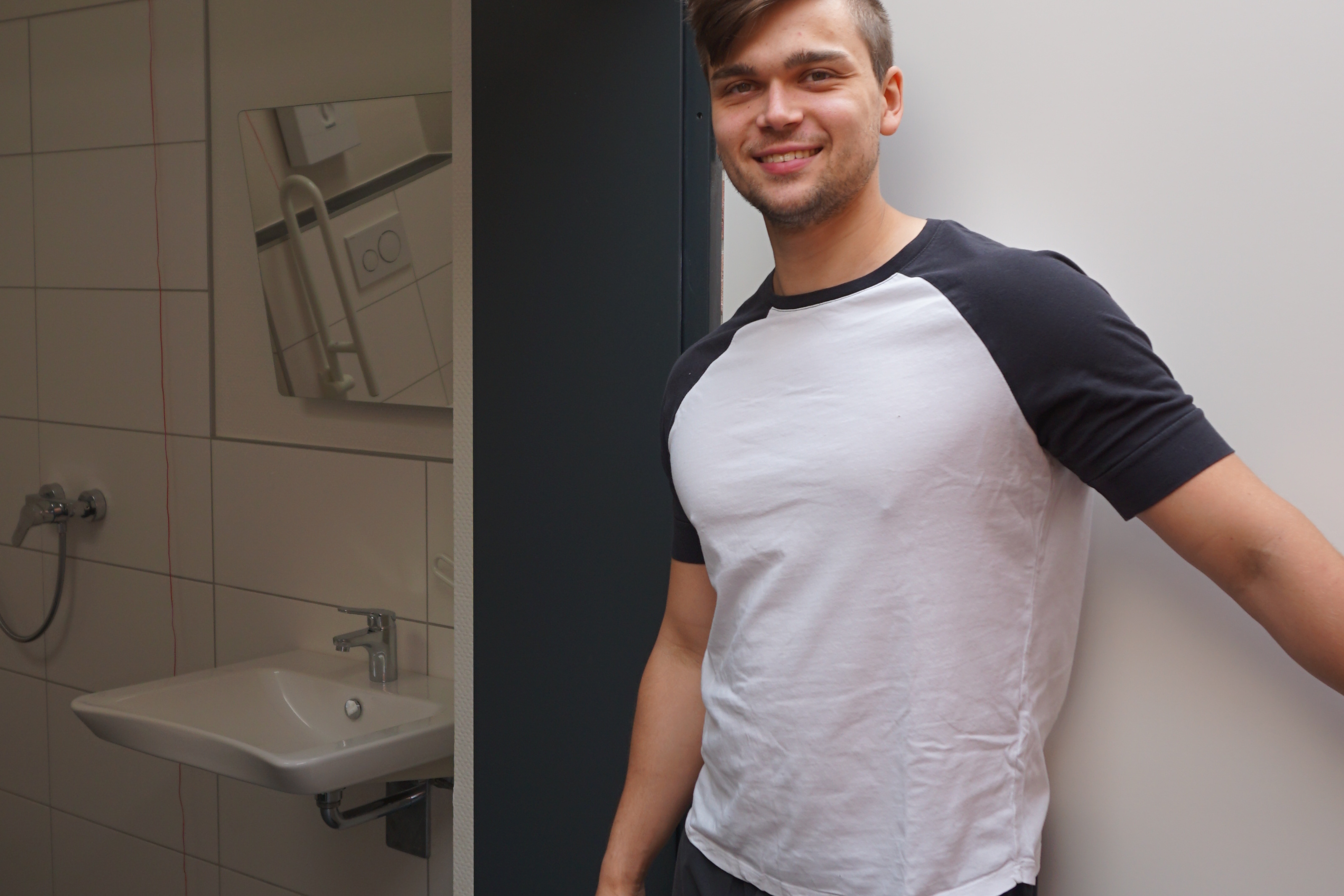 Mann steht vor einer offenen T&uuml;r mit Blick auf ein Badezimmer mit wei&szlig;er Keramik und grauen Fliesen.