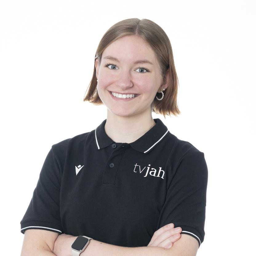 Lächelnde junge Frau in schwarzem Poloshirt mit Logo, die mit verschränkten Armen vor weißem Hintergrund steht.