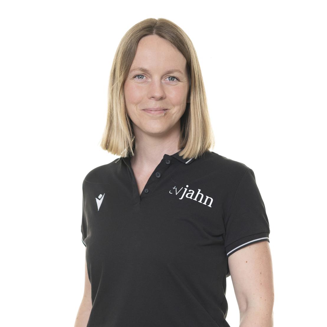 Porträt einer lächelnden Frau mit langen blonden Haaren, in einem schwarzen Poloshirt mit Logo.
