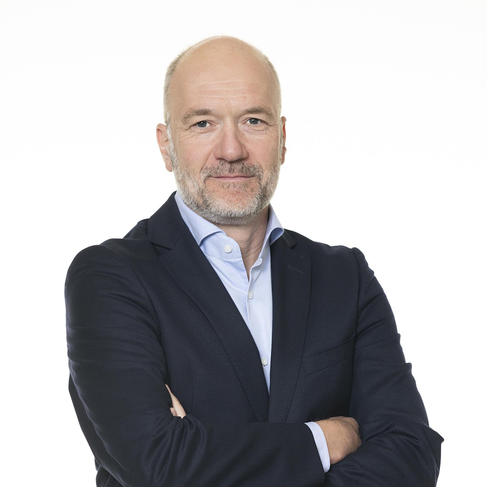 Mann mit grauem Bart und Glatze, tr&auml;gt einen dunkelblauen Anzug und steht mit verschr&auml;nkten Armen vor wei&szlig;em Hintergrund.