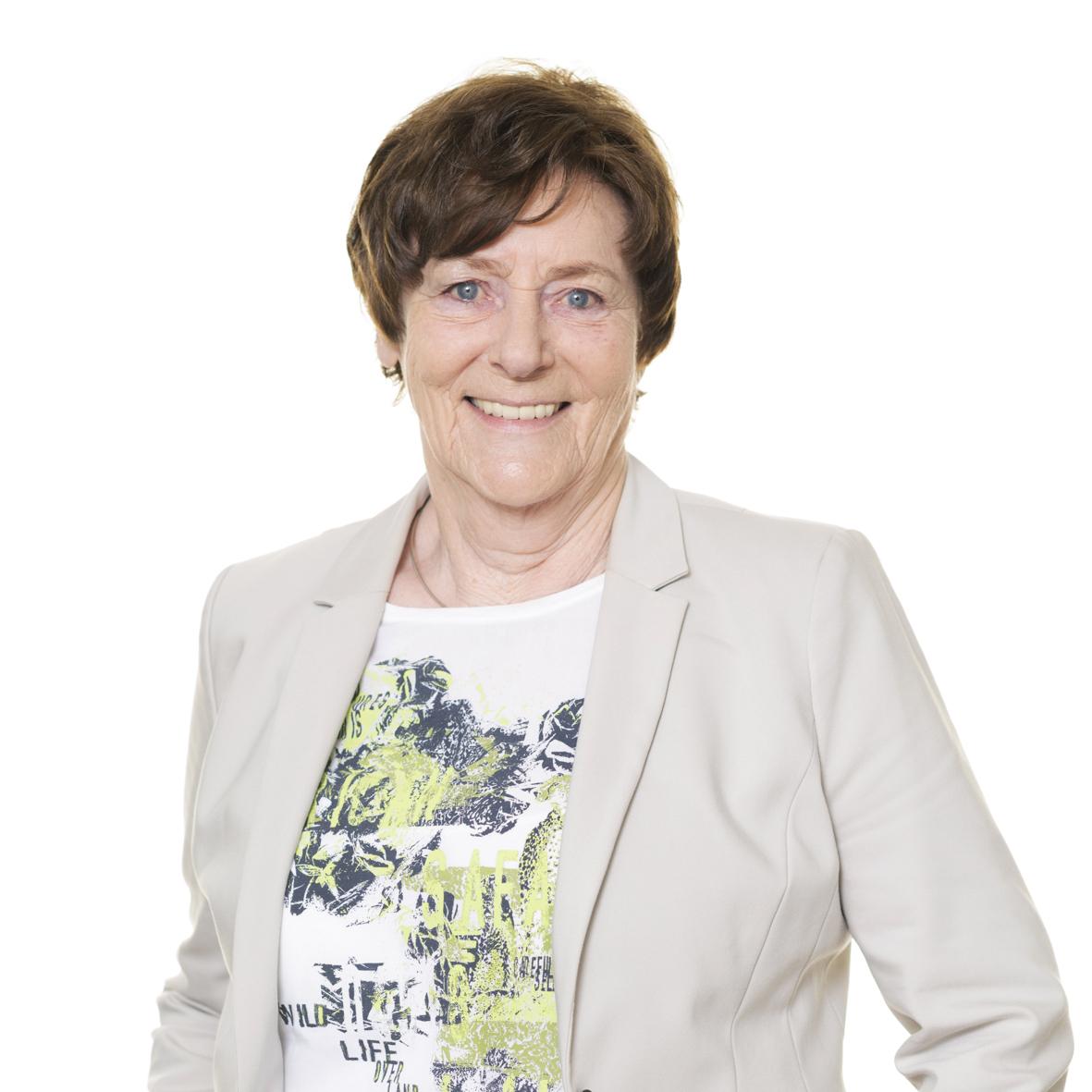 L&auml;chelnde Frau mit kurzen, braunen Haaren und beiger Jacke vor wei&szlig;em Hintergrund. Sie tr&auml;gt ein T-Shirt mit buntem Muster.