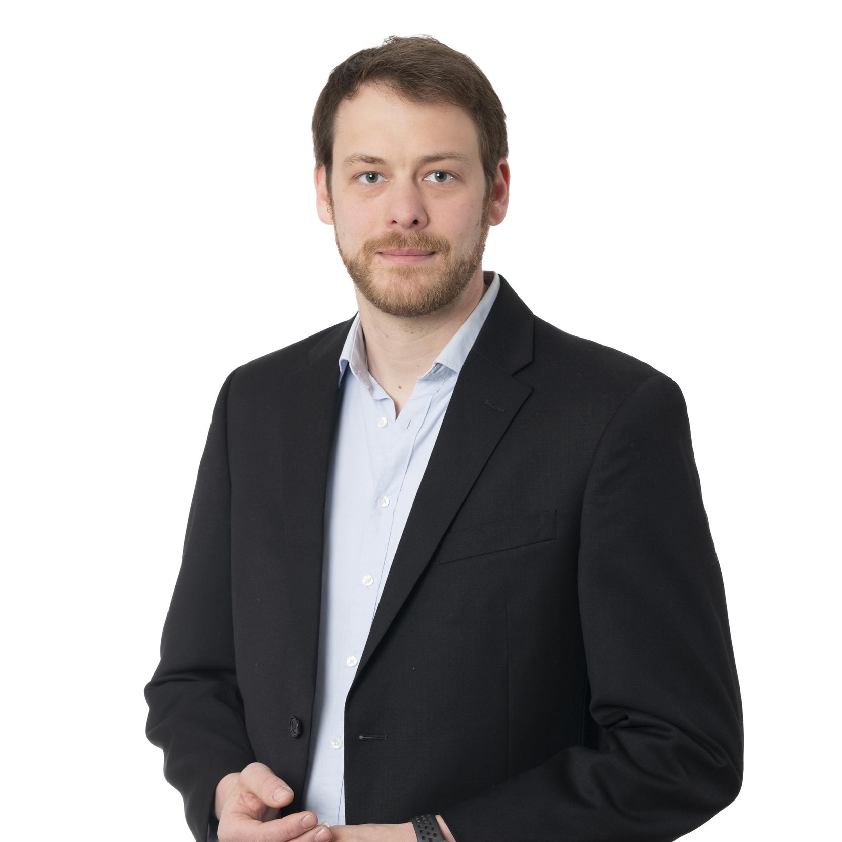 Ein Mann in schwarzem Blazer und hellblauem Hemd steht vor einem wei&szlig;en Hintergrund und schaut direkt in die Kamera.