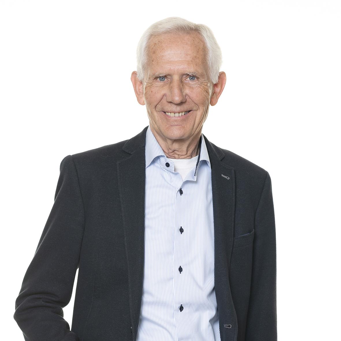 Portr&auml;t eines l&auml;chelnden &auml;lteren Mannes in einem schwarzen Blazer und hellblauen Hemd vor neutralem Hintergrund.