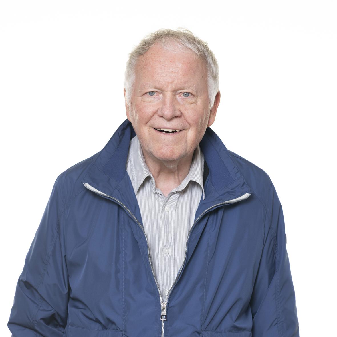 &Auml;lterer Mann mit l&auml;chelndem Gesicht, tr&auml;gt eine blaue Jacke und ein graues Hemd, vor neutralem wei&szlig;en Hintergrund.