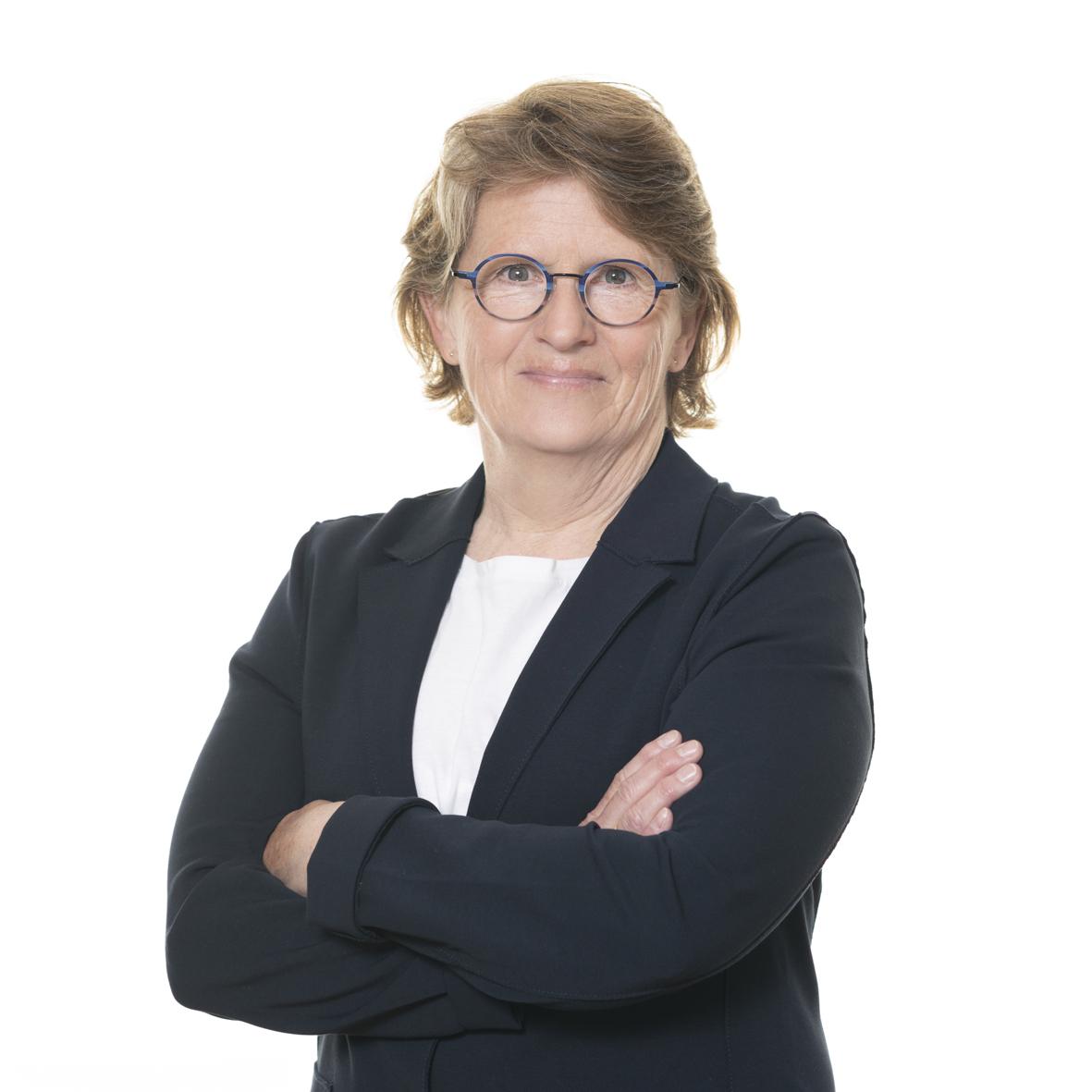 Portr&auml;t einer l&auml;chelnden Frau mit Brille, die in einem schwarzen Blazer mit verschr&auml;nkten Armen steht.