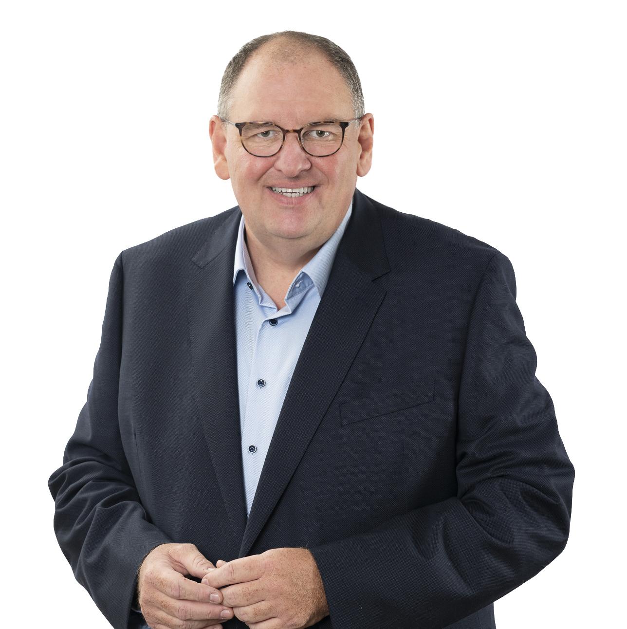 Mann in Brille und Anzug, l&auml;chelnd, mit verschr&auml;nkten H&auml;nden vor wei&szlig;em Hintergrund.