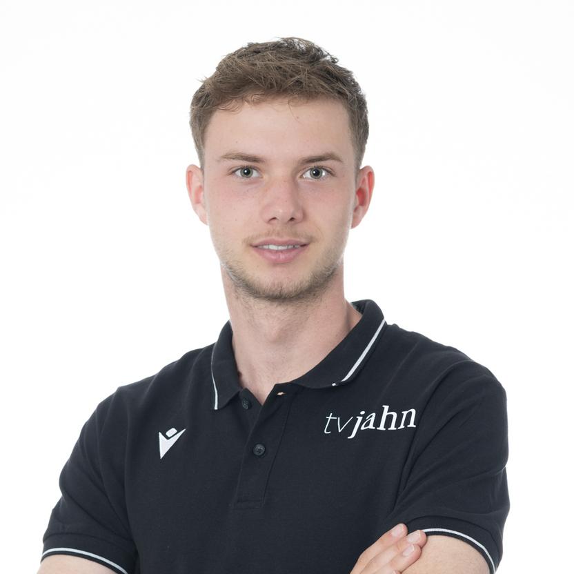 Junger Mann in schwarzem Poloshirt mit Logo, lächelt und hat die Arme vor der Brust verschränkt, vor weißem Hintergrund.