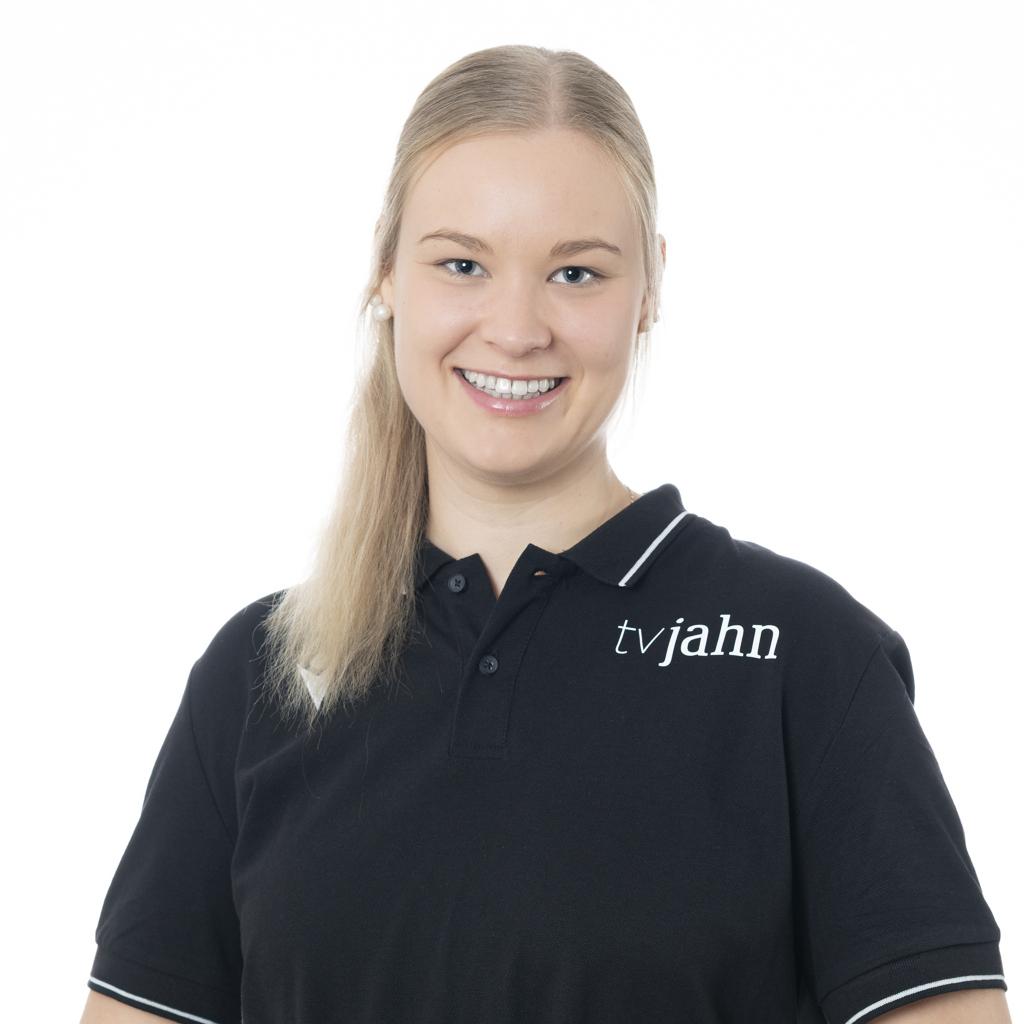 Lächelnde Frau in schwarzem Poloshirt mit Logo „tvjahn“, steht vor hellem Hintergrund und hält eine Hand an ihrem Oberkörper.